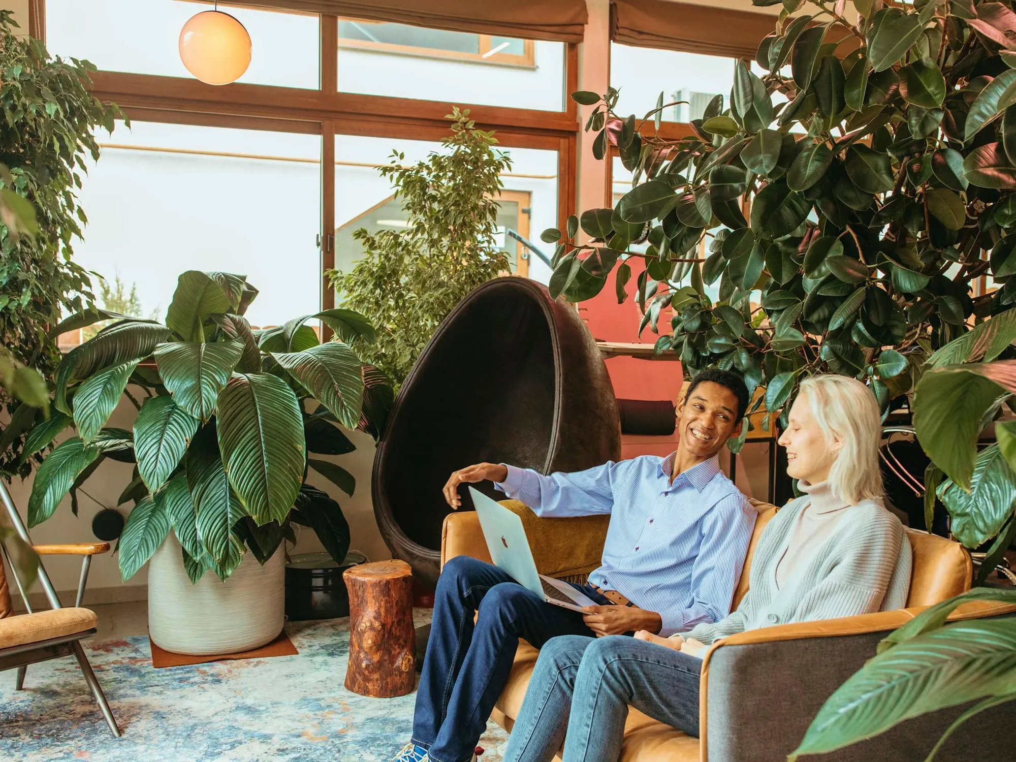 Dos personas sentadas en un sofá rodeado de plantas, conversando con un portátil abierto.